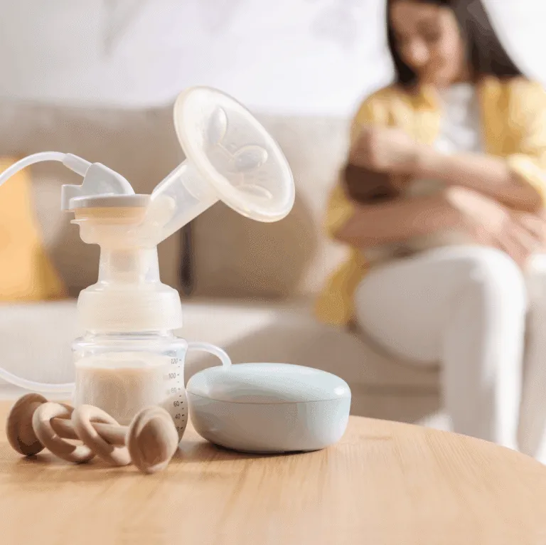 Breast pump and baby bottle on a wooden table, with a mother holding her baby on a sofa in the background
