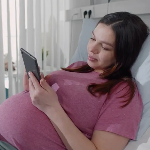 Pregnant woman in a pink top resting in a hospital bed, looking at her phone