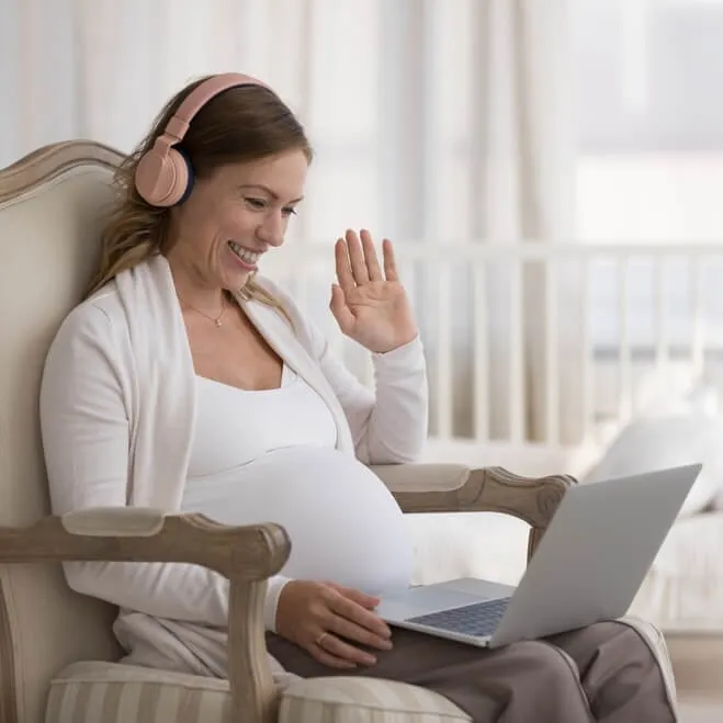 Smiling pregnant woman wearing headphones, waving at a laptop during a video call