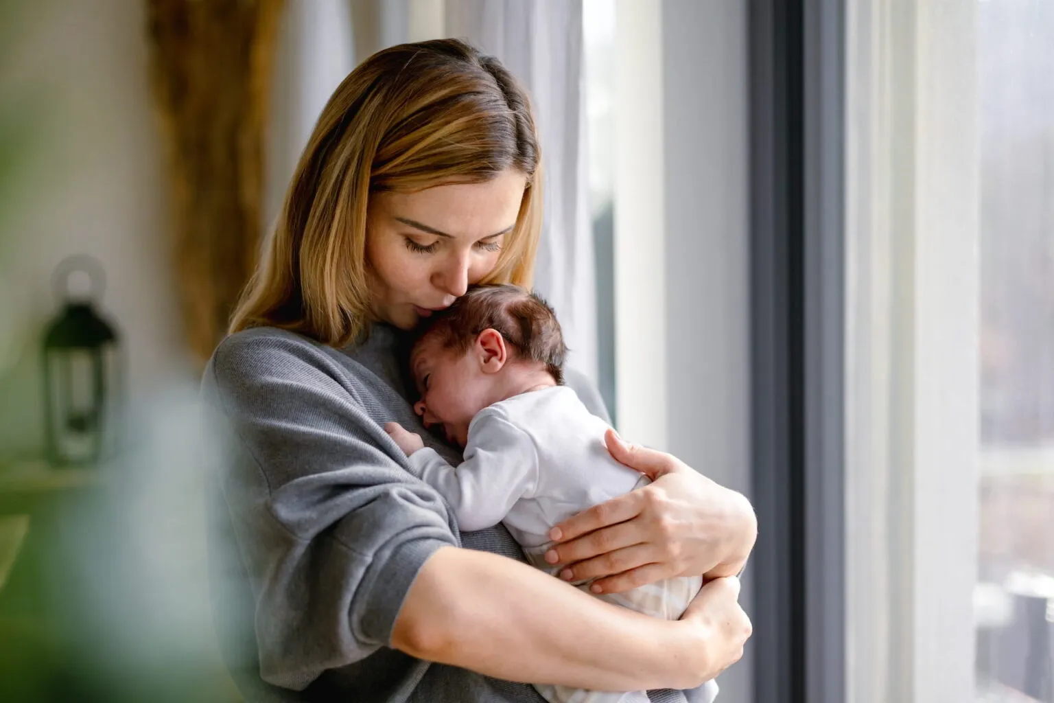 Mother kissing the head of her newborn baby by a window