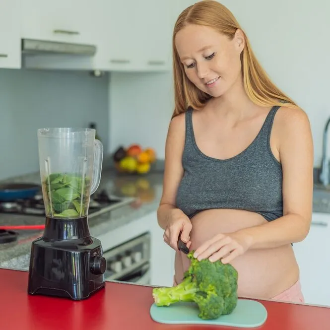 Pregnant woman chopping broccoli beside a blender of green smoothie in her kitchen