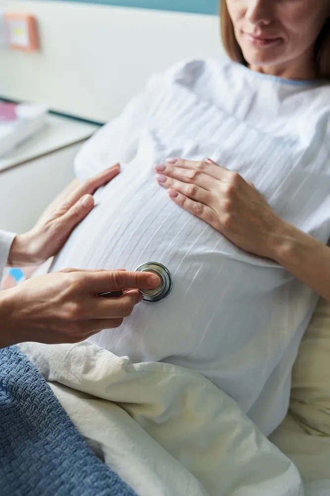 Pregnant woman lying in a hospital bed while a clinician listens to her belly with a stethoscope