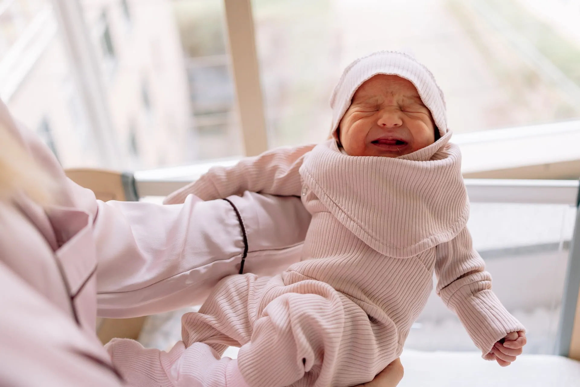 Crying newborn in a pink knit outfit and hat being lifted by a parent