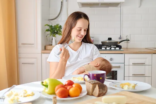 Smiling mother eating a healthy meal while holding her baby at the kitchen table